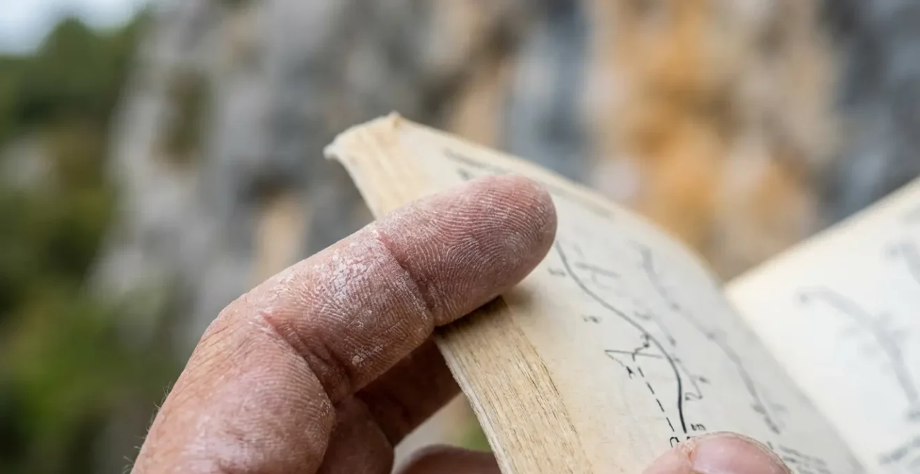 Main tenant un topo d'escalade ouvert avec schémas de voies et cotations visibles en arrière-plan flou de falaise